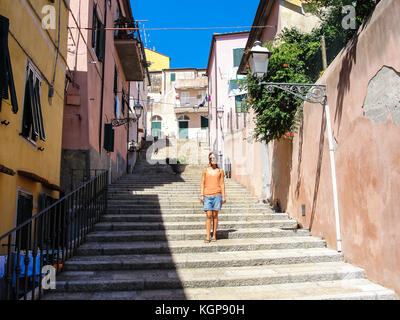 Young woman with sunglasses dressed in casual summer clothes walking down an alley in a small rural medieval village in Italy Stock Photo