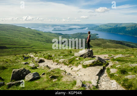 Female hiker admiring the landscape on a path leading to the top of Ben Lomond in a sunny  day. Loch Lomond in the background. Scotland (UK).