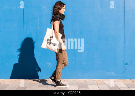 Young girl dressed in casual brown chinos, black sneakers and bag saying 'We love Brixton' walking on a street with blue wall in background. Stock Photo