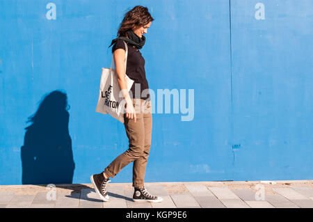 Young girl dressed in casual brown chinos, black sneakers and T-shirt walking on a street with blue wall in background. Stock Photo