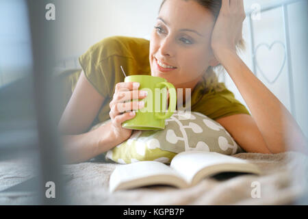 Cheerful woman reading book and drinking tea laying in sofa Stock Photo