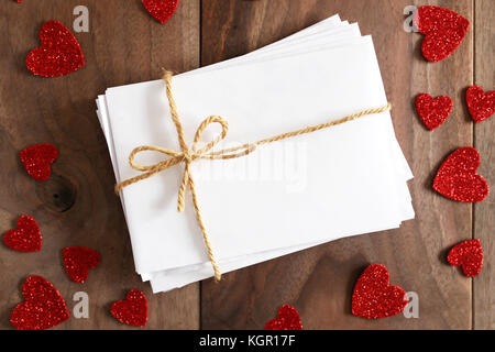 A stack of love letters in blank white envelopes tied with twine string bow is sitting on a rustic cherry wood plank table, surrounded by valentine he Stock Photo
