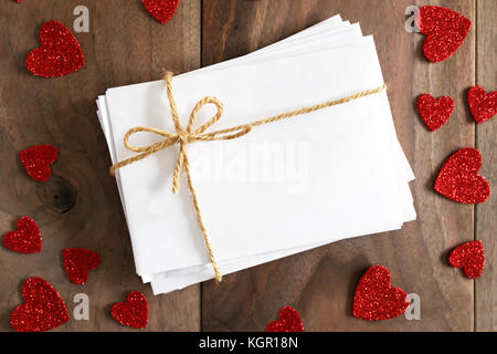 A stack of love letters in blank white envelopes tied with twine string bow is sitting on a rustic cherry wood plank table, surrounded by valentine he Stock Photo