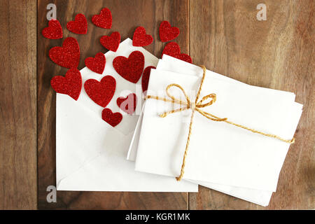 A stack of blank envelopes ties with a twine string bow, are next to an open Valentine's day love letter with red hearts flying out of it, on a rustic Stock Photo