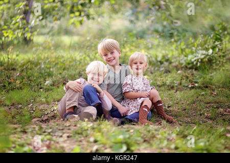 A portrait in the forest of three happy, smiling young children, an eight year old boy, his baby sister and his 5 year old little brother. Stock Photo
