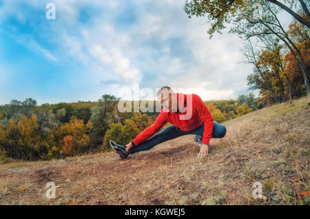 Slim caucasian jogger man in red jacket and black sports leggins performs warming-up  before jogging on the autumnal forest hil with sunset dramatic s Stock Photo