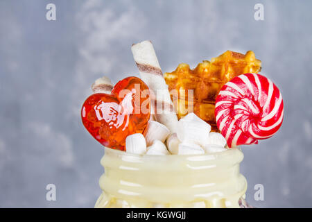 Freakshake from pink smoothie, cream. Monstershake with lollipops, waffles and marshmallow. Extreme milkshake in a Mason jar. Gray dark concrete backg Stock Photo