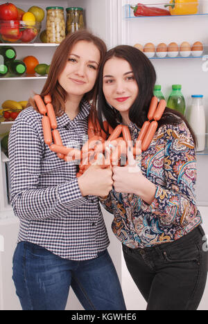 Two young girls holds sausages on the refrigerator background. Two beautiful young girls near the fridge. Stock Photo