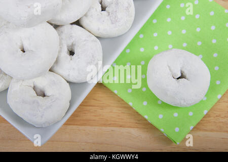 cropped angle view of a square plate with round powdered donuts on a light wood table, one donut isolated on green cloth napkin with white polka dots Stock Photo