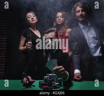 a bearded man with a cigar and cards sitting at a poker table, covered with a green cloth in a club of smoke. Stock Photo