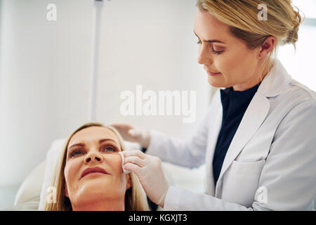 Young female doctor preparing the cheek of a mature woman lying on a table in a beauty clinic for a botox injection Stock Photo