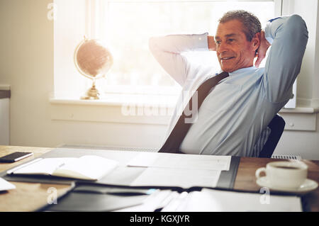 Mature executive in a shirt and tie leaning contently back in his chair with his hands behind his head at his office desk Stock Photo