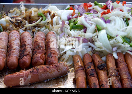 Hot dogs wrapped with thin bacon surrounded by sliced onions, bell peppers, grilling. Street vendor cooking Stock Photo