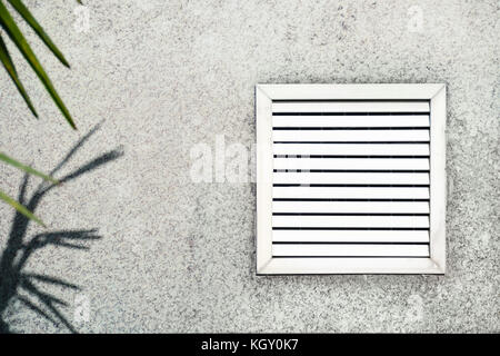 Aged vent with white shutters on the background of gray concrete under the leaves of the palm. Stock Photo