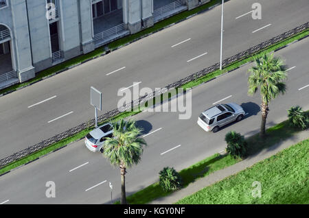 Cars go by road in the city street divided by green spaces and palm trees. View from above. Stock Photo