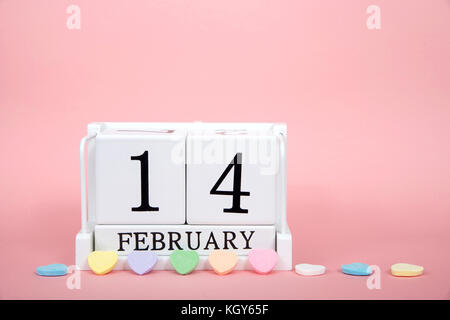 Wood block showing calendar date February 14th, Valentines Day, sitting on pink background with candy hearts lined up in front. Stock Photo