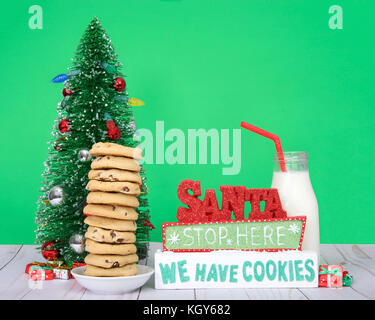 Chocolate chip cookies stacked 12 high on a small round plate, Christmas tree glass bottle of milk with straw next to sign SANTA STOP HERE WE HAVE COO Stock Photo
