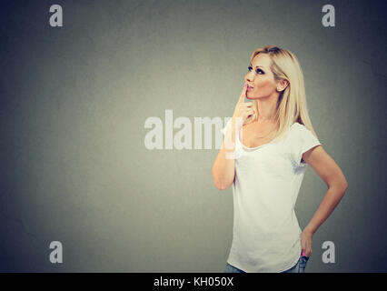 Side profile of a happy woman thinking with finger on chin gesture looking up isolated on gray wall background with copy space. Positive human emotion Stock Photo