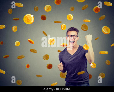Portrait of a happy young man celebrating financial success under a bitcoin rain on a gray wall background. Stock Photo