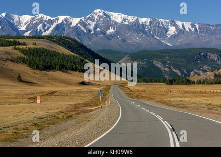Scenic view with the asphalt road in the steppe going to the mountains with snowy peaks and slopes covered with forest on a sunny day Stock Photo