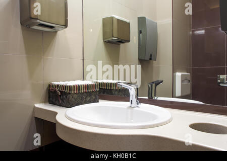 A close-up of a modern white sink, next to it there is a box with cleaned hand towels, automatic hand dryer, in the background a toilet in the toilet  Stock Photo