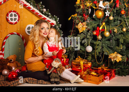 Family Christmas. Mother and daughter are sitting on a blanket at the Christmas tree. The daughter kisses her mother on the cheek. Family holiday. Stock Photo