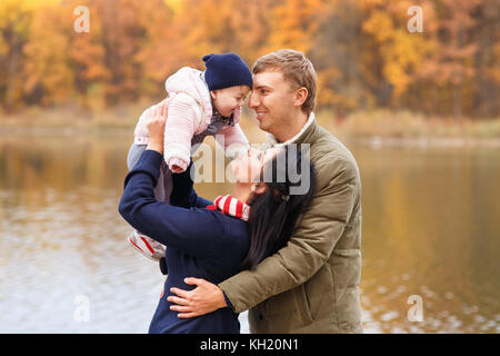 young parents play with little daughter in autumn park near the lake. Mom holding baby girl in hands. Happy family, parental love, autumn season conce Stock Photo