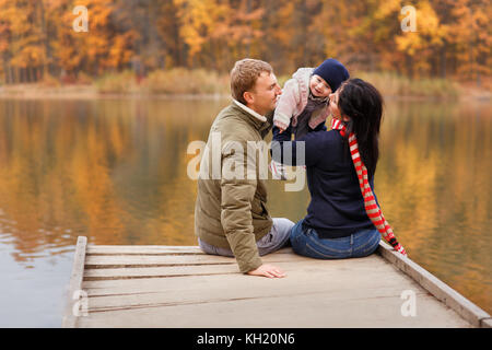 young parents sitting on wooden bridge and play with little daughter autumn forest and river on the background. Mom holding baby girl in hands. Happy  Stock Photo