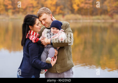 young parents with little daughter in autumn forest near the river. Mom and dad holding baby girl in hands. Happy family, parental love, autumn season Stock Photo
