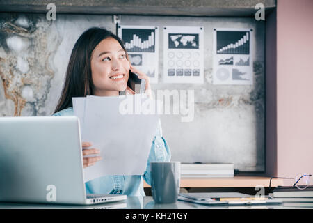 Asian businesswoman talking on mobile phone with customer and looking to work sheet paper in front of laptop computer at office,Office lifestyle conce Stock Photo