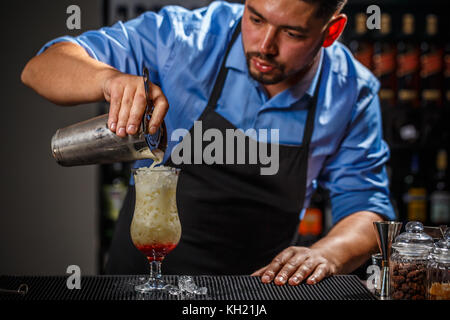 Male bartender is making cocktail pouring alcohol from shaker to glass at bar background Stock Photo