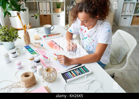 High angle portrait of young female artist painting picture using pastel paints sitting at desk in small art studio Stock Photo