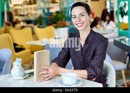 Portrait of modern adult woman smiling happily looking at camera while reading in cafe, copy space Stock Photo