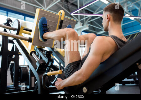 Rear view of muscular sportsman doing leg exercises using machine in modern gym Stock Photo