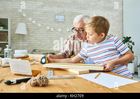 Side view portrait of old man teaching cute little boy woodwork, making wooden models together working at desk in small studio Stock Photo