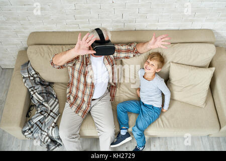 Portrait of excited senior man using VR glasses sitting on sofa at home with laughing grandson beside him Stock Photo