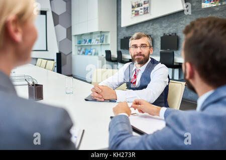 Portrait of barded mature businessman listening  to partners sitting at meeting table in board room while negotiating deal Stock Photo