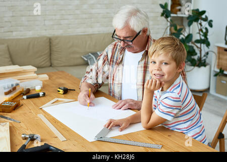 Portrait of cute little boy looking at camera smiling happily while making wooden models with grandpa, sitting at desk in small studio, copy space Stock Photo