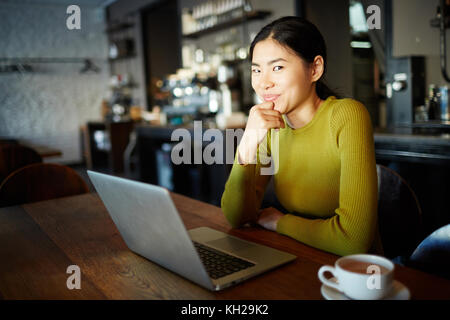 Young Asian girl with laptop watching curious video in cafe by cup of drink
