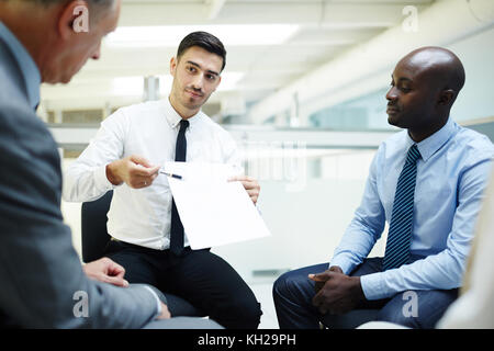 Young economist pointing at contract or financial paper between his two colleagues Stock Photo