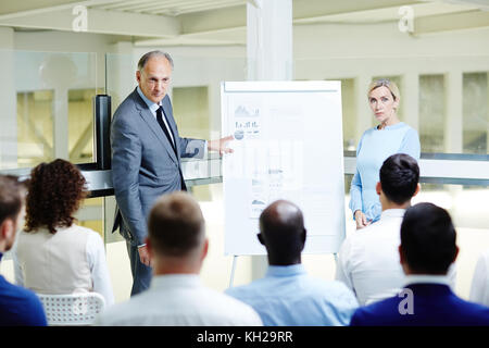 Confident mature expert by whiteboard making presentation of data and explaining it to learners Stock Photo
