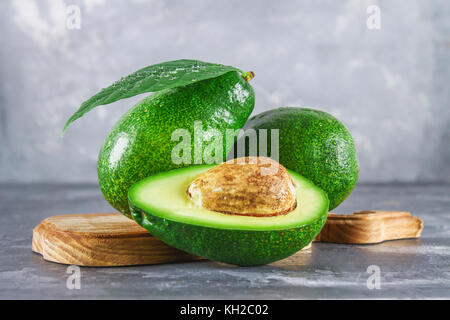 Three green raw ripe avocado fruits and a cut half with a bone with leaves on a wooden cutting board on a gray dark concrete table. Stock Photo