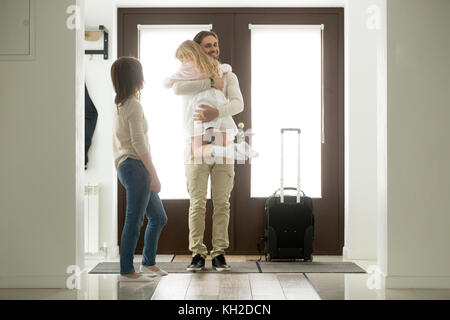 Happy father arrived home returning after business trip with baggage, daddy missed little daughter holding in arms hugging girl while wife standing in