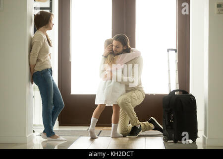 Sad father hugging little daughter before leaving for long business trip, upset dad embracing crying girl saying goodbye to daddy at home in hall with Stock Photo