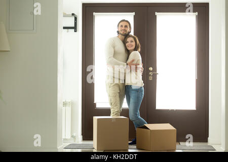 Moving day concept, excited young couple homeowners embracing in new home standing in modern hallway with cardboard boxes on floor looking around, mor