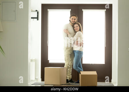 Happy young couple hugging looking at camera moving into new home, smiling homeowners embracing in hallway standing against door near cardboard boxes,