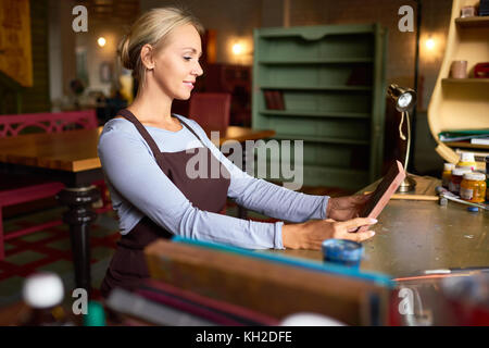 Side view portrait of beautiful young woman enjoying work in art studio holding freshly painted picture and smiling happily Stock Photo