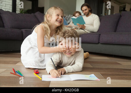 Little sister closing brothers eyes with hands, siblings playing peek a boo laughing on floor, parents reading book on sofa at home, family with child Stock Photo