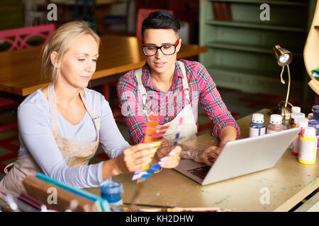 Portrait of two creative young women enjoying  work together in art studio, using laptop and holding swatches while choosing colors for new project Stock Photo