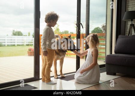 Children caressing watch dog protecting home, kids playing with big pet coming inside house, little boy and girl having fun stroking german shepherd s Stock Photo